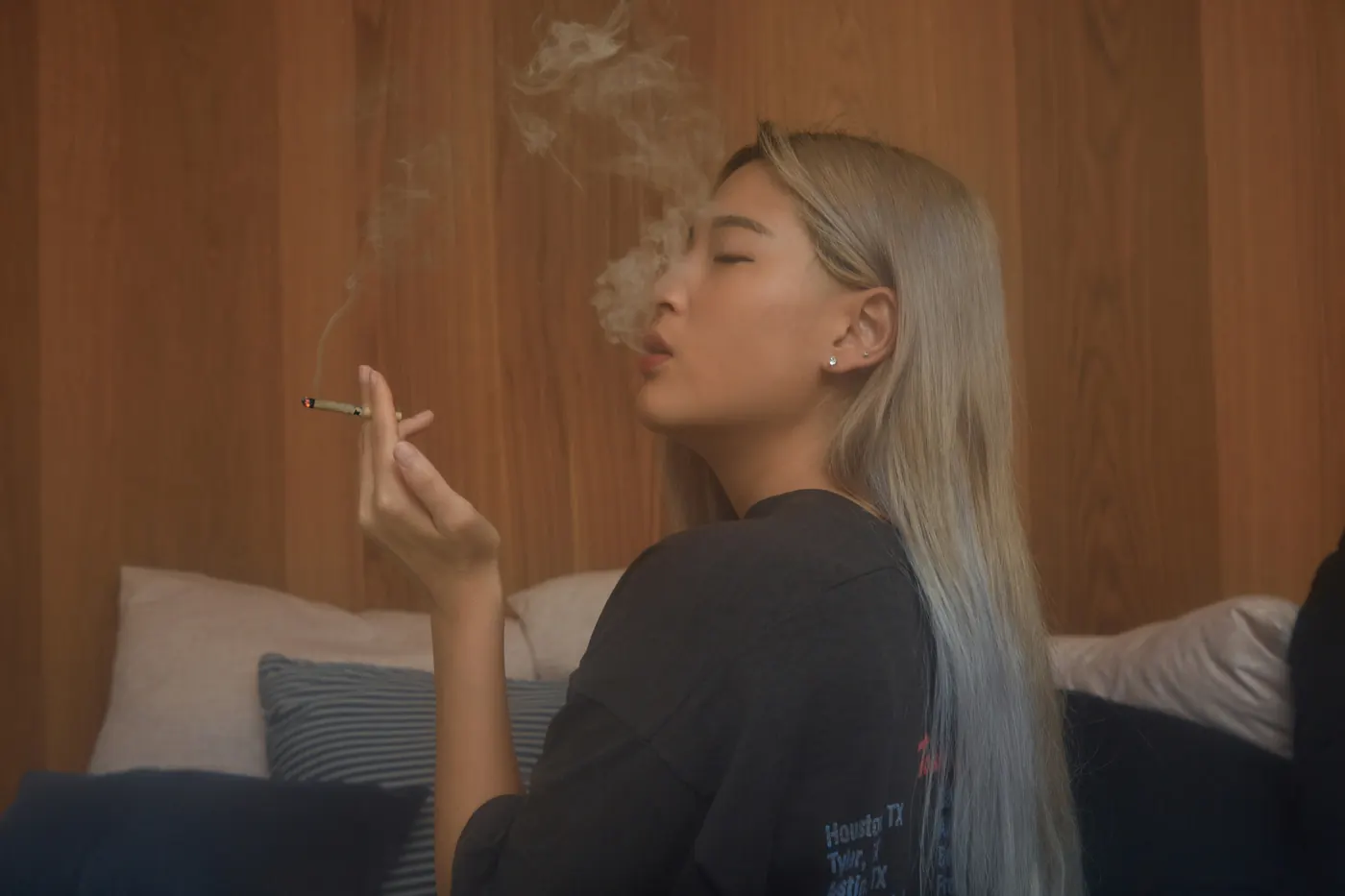 A woman exhaling smoke against a warm wood wall, pre-roll in hand