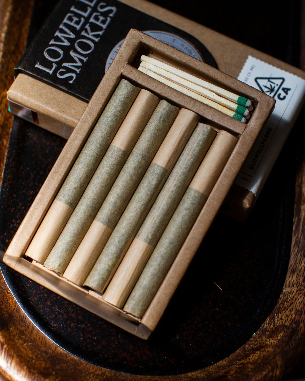 An open Lowell Smokes pack: the tray of six pre-rolls slid out, emergency matches in the top slot, the magnetic flap behind